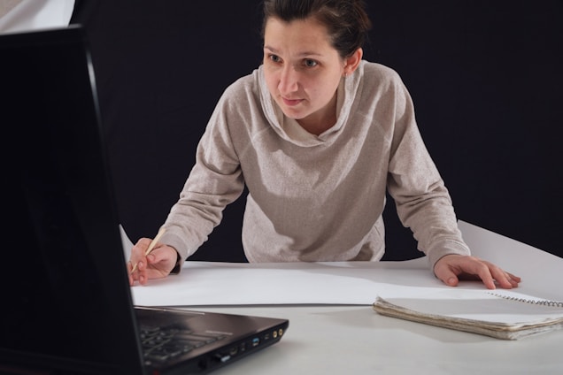 A person leaning over a table, focused on drawing or writing on a large sheet of paper while looking at the screen of an open laptop. Their attire is casual, and they hold a pencil in their right hand. A notebook is placed on the table beside them.