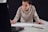 A person leaning over a table, focused on drawing or writing on a large sheet of paper while looking at the screen of an open laptop. Their attire is casual, and they hold a pencil in their right hand. A notebook is placed on the table beside them.