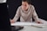 A person leaning over a table, focused on drawing or writing on a large sheet of paper while looking at the screen of an open laptop. Their attire is casual, and they hold a pencil in their right hand. A notebook is placed on the table beside them.