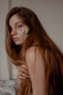 A person with long brown hair is looking over their shoulder, displaying a calm and introspective expression. There is an artistic floral design painted on their cheek, accentuating their serene demeanor.