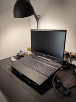 A workspace features a laptop on a dark desk with a sleek lamp overhead. There are additional items such as a cable and electronic gadgets nearby, and a small plant adds a touch of greenery.