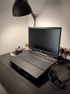 A workspace features a laptop on a dark desk with a sleek lamp overhead. There are additional items such as a cable and electronic gadgets nearby, and a small plant adds a touch of greenery.