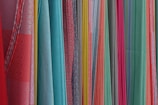 A variety of colorful fabrics are arranged vertically, showcasing different textures and patterns. The fabrics range in color from bright reds, pinks, and yellows to light blues and greens. Some fabrics have intricate geometric patterns, while others have solid colors or stripes.