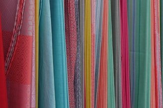 A variety of colorful fabrics are arranged vertically, showcasing different textures and patterns. The fabrics range in color from bright reds, pinks, and yellows to light blues and greens. Some fabrics have intricate geometric patterns, while others have solid colors or stripes.