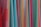 A variety of colorful fabrics are arranged vertically, showcasing different textures and patterns. The fabrics range in color from bright reds, pinks, and yellows to light blues and greens. Some fabrics have intricate geometric patterns, while others have solid colors or stripes.
