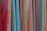 A variety of colorful fabrics are arranged vertically, showcasing different textures and patterns. The fabrics range in color from bright reds, pinks, and yellows to light blues and greens. Some fabrics have intricate geometric patterns, while others have solid colors or stripes.