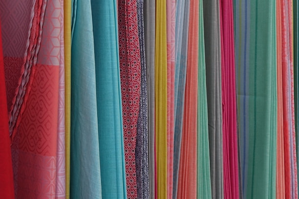 A variety of colorful fabrics are arranged vertically, showcasing different textures and patterns. The fabrics range in color from bright reds, pinks, and yellows to light blues and greens. Some fabrics have intricate geometric patterns, while others have solid colors or stripes.