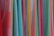 A variety of colorful fabrics are arranged vertically, showcasing different textures and patterns. The fabrics range in color from bright reds, pinks, and yellows to light blues and greens. Some fabrics have intricate geometric patterns, while others have solid colors or stripes.