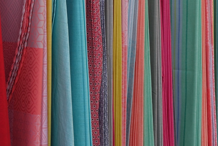 A variety of colorful fabrics are arranged vertically, showcasing different textures and patterns. The fabrics range in color from bright reds, pinks, and yellows to light blues and greens. Some fabrics have intricate geometric patterns, while others have solid colors or stripes.