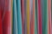 A variety of colorful fabrics are arranged vertically, showcasing different textures and patterns. The fabrics range in color from bright reds, pinks, and yellows to light blues and greens. Some fabrics have intricate geometric patterns, while others have solid colors or stripes.