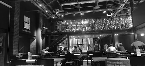 A black and white image of a bar interior with exposed industrial-style ceilings, decorative lights hanging from a railing, and people seated at tables. A person is playing guitar to the left. The setting has a warm and inviting atmosphere, with visible branding on the walls indicating a sense of local identity.
