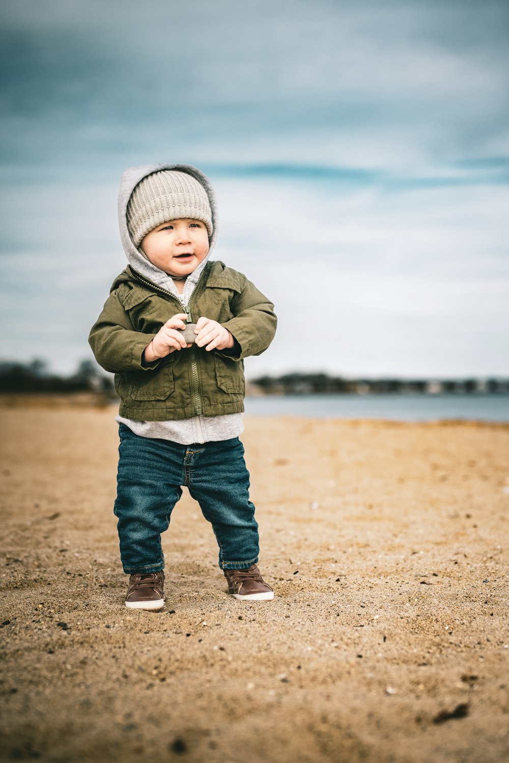 a toddler standing on a beach wearing a jacket and hat