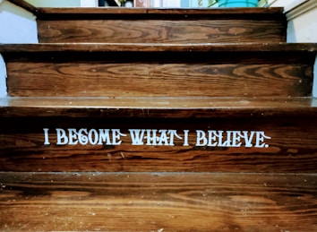 Wooden steps with a motivational quote displayed in bold white letters reading 'I BECOME WHAT I BELIEVE.' The texture of the wood grain is visible, and there is a dim lighting atmosphere.