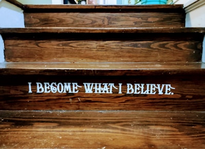 Wooden steps with a motivational quote displayed in bold white letters reading 'I BECOME WHAT I BELIEVE.' The texture of the wood grain is visible, and there is a dim lighting atmosphere.