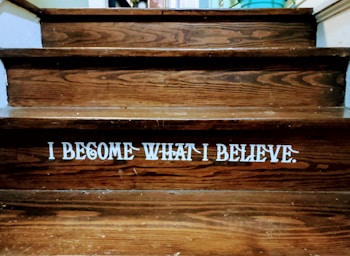 Wooden steps with a motivational quote displayed in bold white letters reading 'I BECOME WHAT I BELIEVE.' The texture of the wood grain is visible, and there is a dim lighting atmosphere.