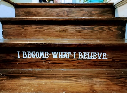 Wooden steps with a motivational quote displayed in bold white letters reading 'I BECOME WHAT I BELIEVE.' The texture of the wood grain is visible, and there is a dim lighting atmosphere.