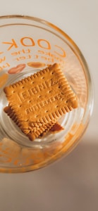 A square butter biscuit is placed inside a clear glass. The glass has orange text and a heart illustration on it. The biscuit is labeled 'LEIBNIZ BUTTERKEKS'.