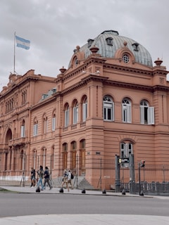 A historic, neoclassical building with a pink façade and a distinct domed roof. The structure is adorned with ornate architectural details and large windows, and the Argentine flag is flying at the top. A fenced area surrounds the building. Several people are walking along the sidewalk in front of the building.