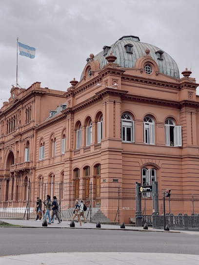 A historic, neoclassical building with a pink façade and a distinct domed roof. The structure is adorned with ornate architectural details and large windows, and the Argentine flag is flying at the top. A fenced area surrounds the building. Several people are walking along the sidewalk in front of the building.
