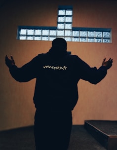 A person stands with outstretched arms in front of a cross-shaped window made of frosted glass blocks. The person's back is facing the viewer, and they are wearing a dark hoodie with a website address printed on the back. The room is dimly lit, creating a contemplative atmosphere.