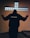 A person stands with outstretched arms in front of a cross-shaped window made of frosted glass blocks. The person's back is facing the viewer, and they are wearing a dark hoodie with a website address printed on the back. The room is dimly lit, creating a contemplative atmosphere.
