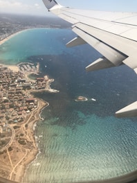 An aerial view of a coastal region with clear blue water and visible land formations. The coastline is lined with buildings and a marina, while the airplane wing is prominently visible on the right side of the image.