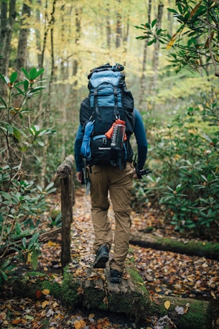 A person with a large backpack is walking along a narrow wooden bridge in a forest. The surroundings are lush with greenery and autumn leaves scattered on the ground. The person appears to be hiking, equipped with outdoor gear including climbing rope and a water bottle attached to the backpack.