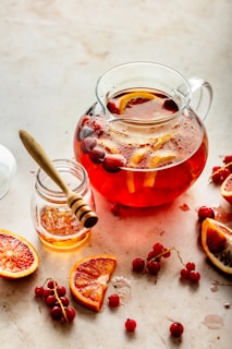 A glass jug filled with a red liquid, possibly a fruit-infused drink, containing slices of citrus and whole cranberries. Beside the jug, there is a small jar of honey with a wooden honey dipper resting inside. Surrounding the jug are halved blood oranges and loose cranberries scattered on a light-colored surface.