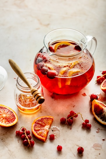 A glass jug filled with a red liquid, possibly a fruit-infused drink, containing slices of citrus and whole cranberries. Beside the jug, there is a small jar of honey with a wooden honey dipper resting inside. Surrounding the jug are halved blood oranges and loose cranberries scattered on a light-colored surface.