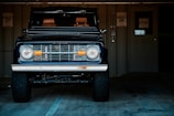 A classic off-road vehicle with a vintage style front grille and headlights is parked inside a dimly lit garage. The vehicle has large, rugged tires and a dark, sleek paint finish. Behind it, a metal wall with a door and an electrical fixture is visible, along with some parking signs.