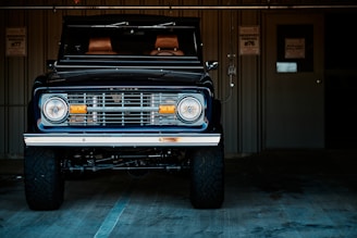 A classic off-road vehicle with a vintage style front grille and headlights is parked inside a dimly lit garage. The vehicle has large, rugged tires and a dark, sleek paint finish. Behind it, a metal wall with a door and an electrical fixture is visible, along with some parking signs.