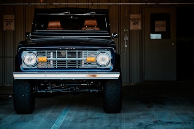A classic off-road vehicle with a vintage style front grille and headlights is parked inside a dimly lit garage. The vehicle has large, rugged tires and a dark, sleek paint finish. Behind it, a metal wall with a door and an electrical fixture is visible, along with some parking signs.
