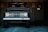 A classic off-road vehicle with a vintage style front grille and headlights is parked inside a dimly lit garage. The vehicle has large, rugged tires and a dark, sleek paint finish. Behind it, a metal wall with a door and an electrical fixture is visible, along with some parking signs.
