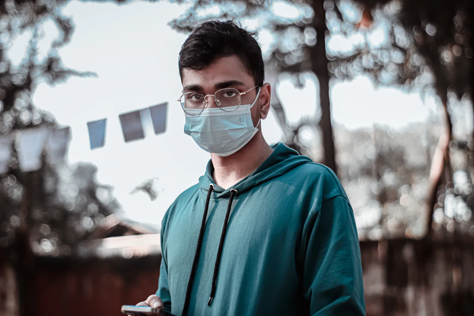 In the image, a young man is standing outdoors, wearing a blue hoodie and a face mask. He holds a cell phone in his hand, which he is using to take a photo or make a call. The background features trees and flags fluttering in the breeze, creating an outdoor setting with a sense of movement and activity.

The young man's attire suggests that it might be a cold day, as he has chosen to wear a face mask for protection against airborne particles or germs. His choice of clothing also indicates that he is dressed casually, possibly for a day out in the park or other outdoor activities.