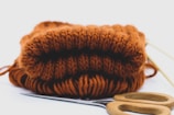 A folded piece of rust-colored knitted fabric is placed on a white surface, accompanied by a pair of wooden-handled scissors and a knitting needle.