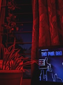 A dark room illuminated with red lighting features a curtain with a floral design, a potted plant with long leaves, and a digital screen displaying a stylized cityscape and text in a graphical design.