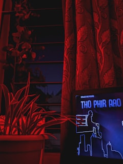 A dark room illuminated with red lighting features a curtain with a floral design, a potted plant with long leaves, and a digital screen displaying a stylized cityscape and text in a graphical design.