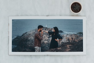 A photo album lies open on a marble surface showing a couple by the rocky seaside. The man, dressed in a plaid shirt and beige pants, gently kisses the woman's hand as she looks on in a black dress, against a backdrop of ocean and rocks. A small potted plant with an aloe vera is positioned near the top right corner of the frame.