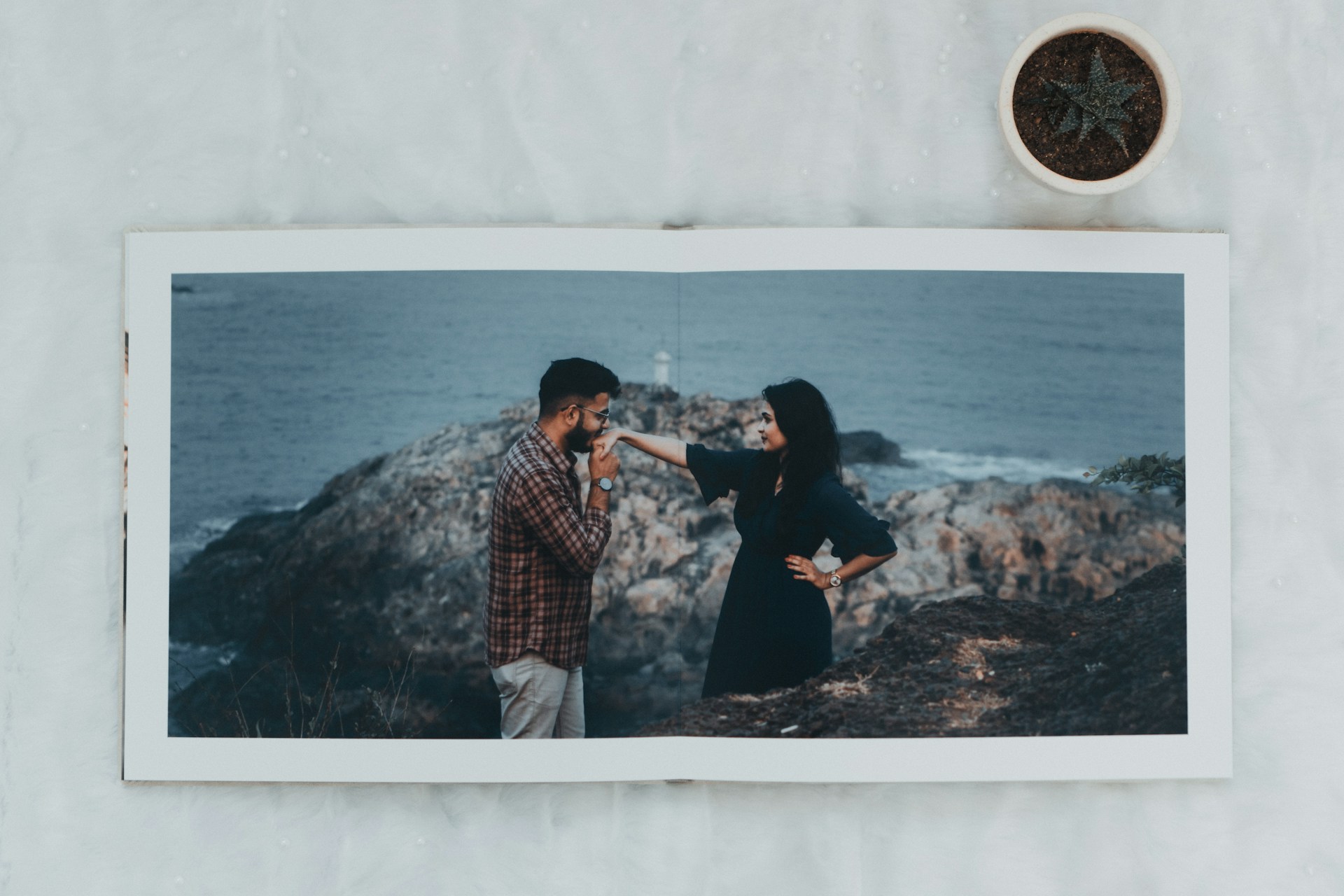 A photo album lies open on a marble surface showing a couple by the rocky seaside. The man, dressed in a plaid shirt and beige pants, gently kisses the woman's hand as she looks on in a black dress, against a backdrop of ocean and rocks. A small potted plant with an aloe vera is positioned near the top right corner of the frame.