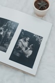 A photo album opened to a page displaying two black and white photographs. The left photo features a man standing by a tree, and the right photo shows a couple dancing under string lights. A small potted succulent is placed next to the album on a soft, white surface.