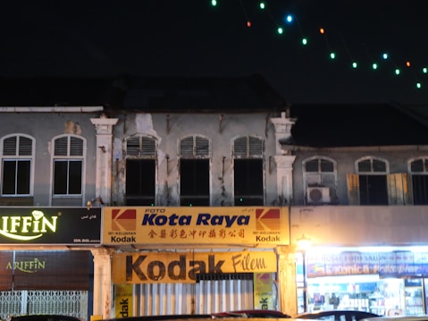 A row of old shophouses at night, with visible signs and logos for Kodak and other businesses. The buildings have an aged appearance, with peeling paint and a classic architectural style featuring arched windows. String lights with multicolored bulbs hang above, adding some brightness to the scene.