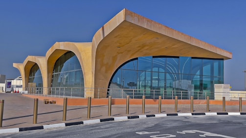 A modern architectural structure features large curved arches and extensive glass windows, creating a sleek and contemporary look. The building sits on a paved area with bollards lining the edge of the walkway. The clear blue sky provides a vibrant backdrop.
