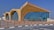A modern architectural structure features large curved arches and extensive glass windows, creating a sleek and contemporary look. The building sits on a paved area with bollards lining the edge of the walkway. The clear blue sky provides a vibrant backdrop.