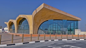A modern architectural structure features large curved arches and extensive glass windows, creating a sleek and contemporary look. The building sits on a paved area with bollards lining the edge of the walkway. The clear blue sky provides a vibrant backdrop.
