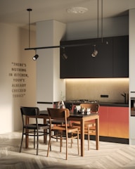 A modern kitchen with a minimalist design features a table and chairs set in a room with light-colored wooden flooring. The cabinetry is mostly dark with a warm gradient. Above the cabinets, there is ambient lighting. A stovetop with a sleek faucet is integrated into the counters. A quote is artistically displayed on the wall.