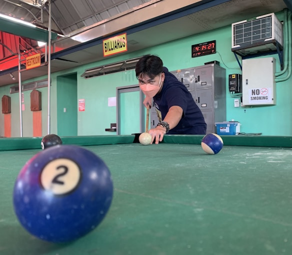 A person wearing a mask is playing billiards indoors, aiming to strike the cue ball with a cue stick. The billiard table is green, and several balls are visible, including a blue ball with the number '2'. The room has signs for 'restroom' and 'billiards', with a clock showing the time and a 'no smoking' sign on the wall.