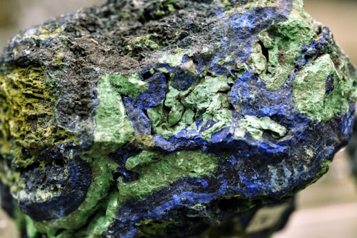 A rough mineral rock with an intricate pattern of green and blue hues, showcasing layers and textures. The surface appears uneven and rugged, with some areas reflecting light, emphasizing the vivid colors.