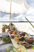 A picnic setup on a beach features an array of colorful fruits, breads, and cheeses arranged on a long wooden board. A white fringed umbrella provides shade, with greenery and decorative elements enhancing the setting. The background reveals a sandy beach extending towards a distant tree-lined shore beneath cloudy skies.