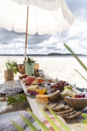 A picnic setup on a beach features an array of colorful fruits, breads, and cheeses arranged on a long wooden board. A white fringed umbrella provides shade, with greenery and decorative elements enhancing the setting. The background reveals a sandy beach extending towards a distant tree-lined shore beneath cloudy skies.