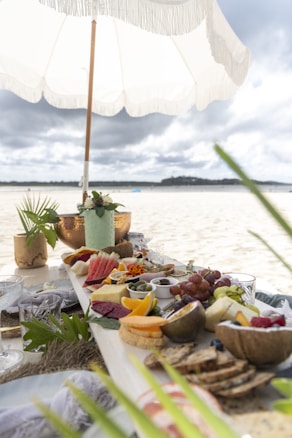 A picnic setup on a beach features an array of colorful fruits, breads, and cheeses arranged on a long wooden board. A white fringed umbrella provides shade, with greenery and decorative elements enhancing the setting. The background reveals a sandy beach extending towards a distant tree-lined shore beneath cloudy skies.